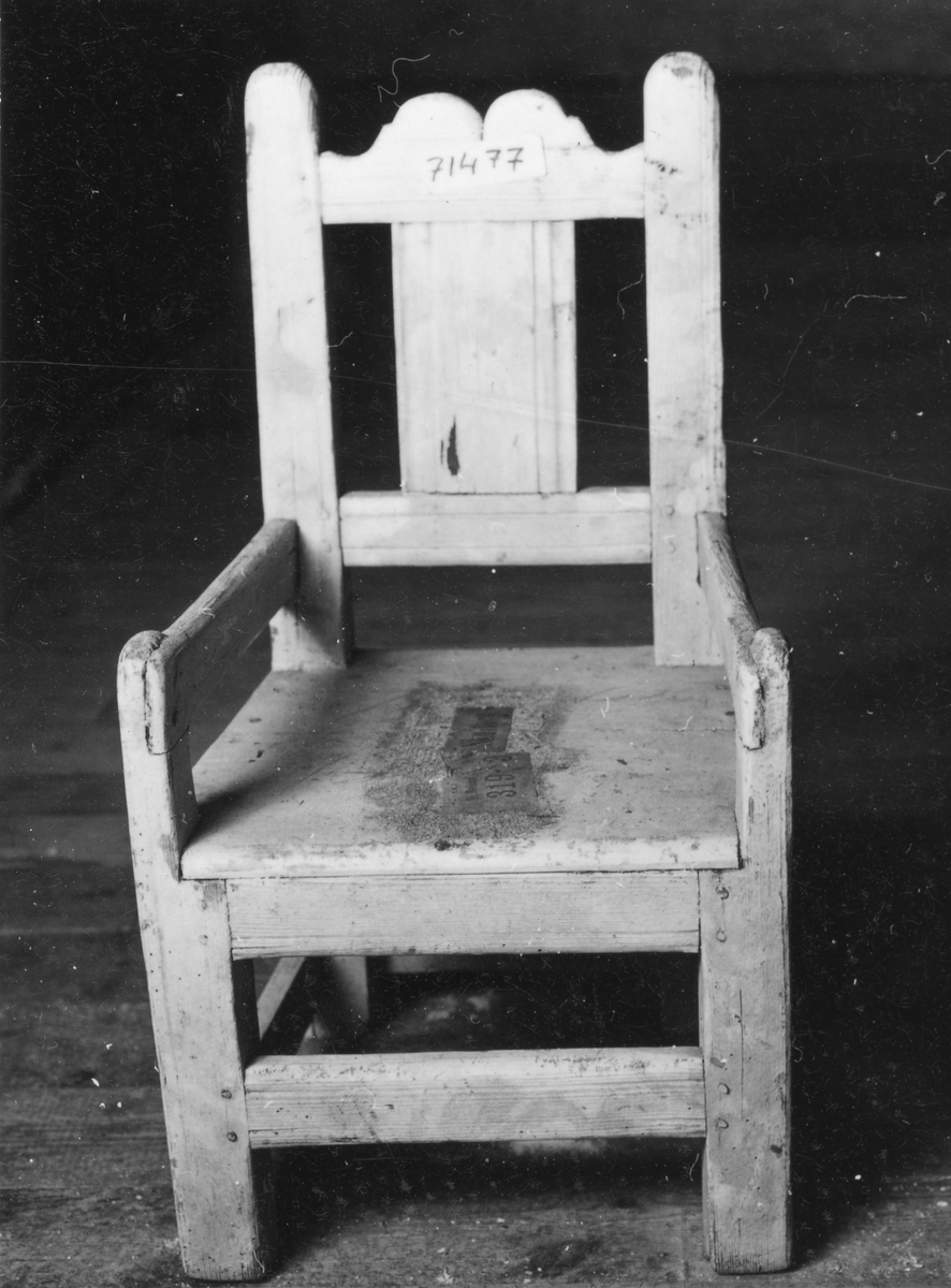 Vitmålad barnstol med vertikal ryggbrick och profilerat krön, raka armstöd fästade i frambenen som skjuter upp ovanför sitsen.
