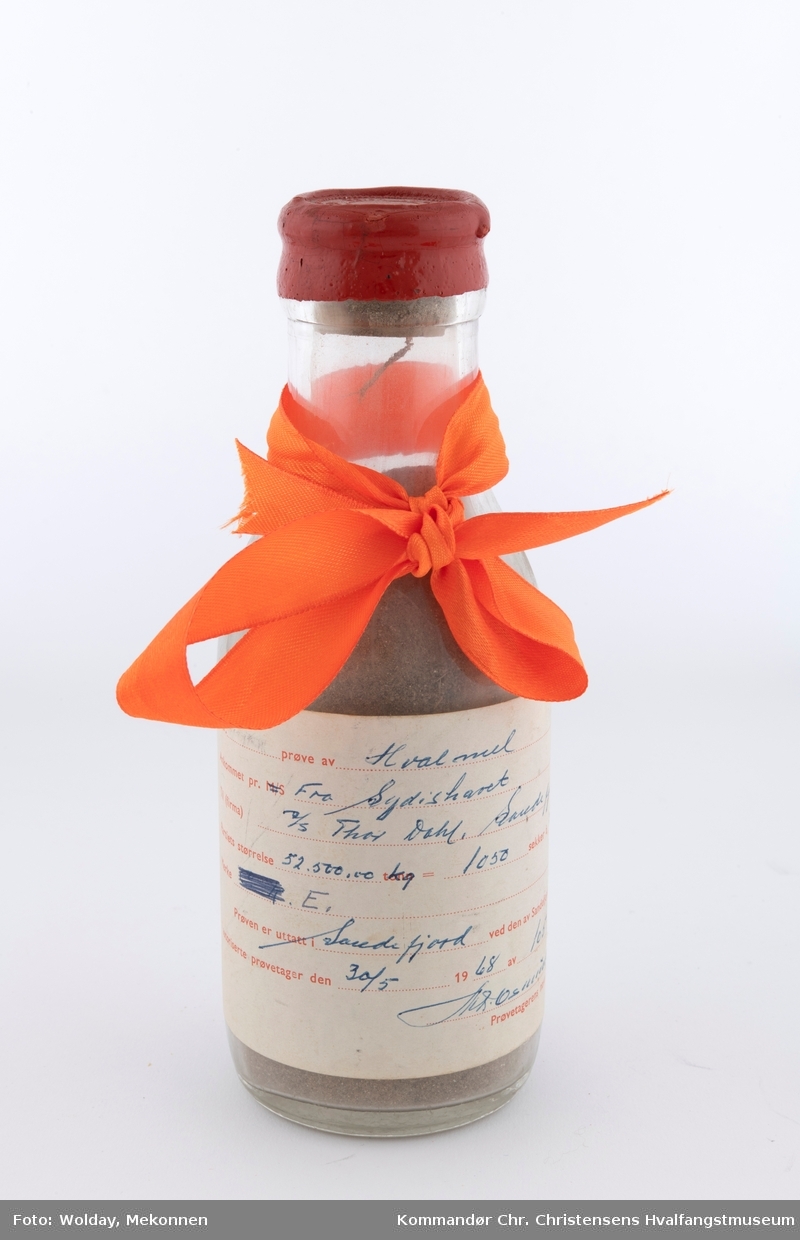 Glassflaske med etikett og vokset kort. Oransje gavebånd rundt flaskehalsen.
Flaske med prøve av hvalmel
