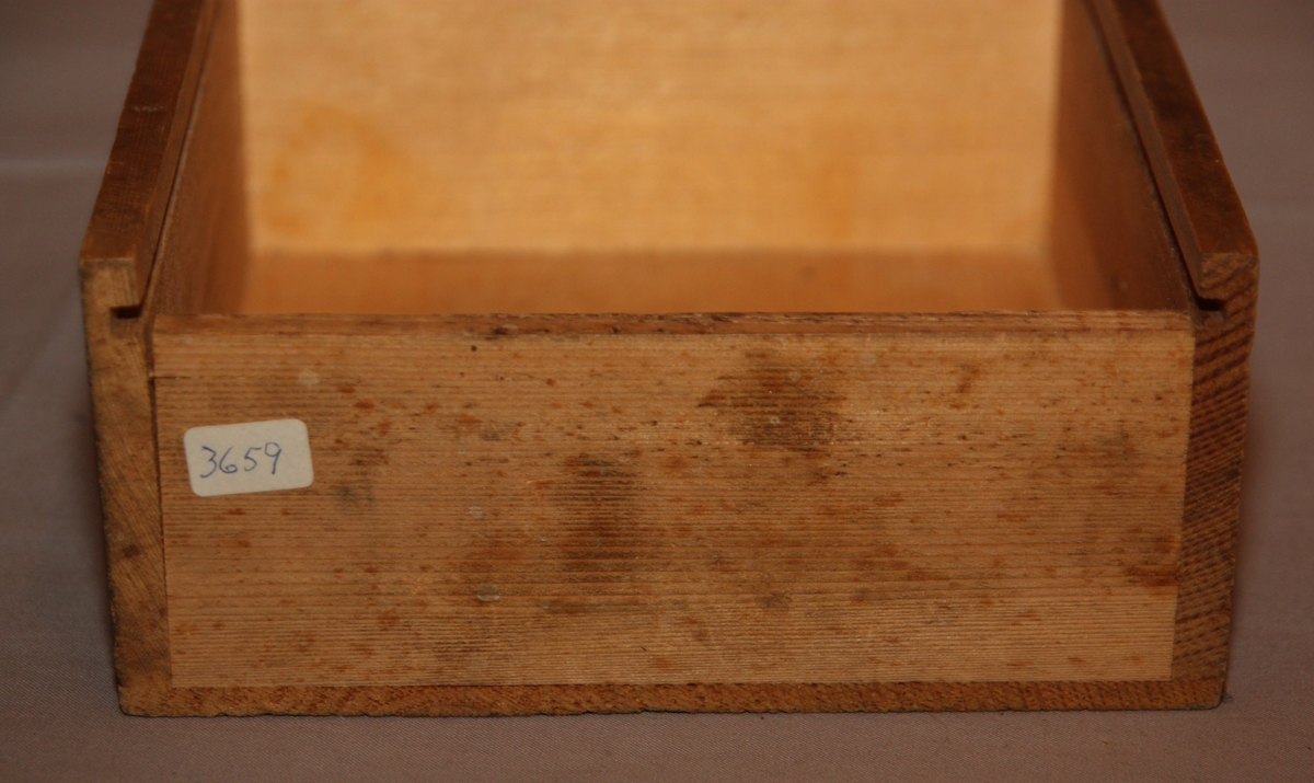 Bygglåda, rektangulär låda av furu med inskjutbart lock från Gemla Leksaksfabriks Aktiebolag (1866-1884). Utan leksaker innuti. Etikett på locket som föreställer en logga med en gunghäst omgiven av tre vita blommor. Etikettens yttre kanter har ett dekorelement föreställande ett slingrande band.