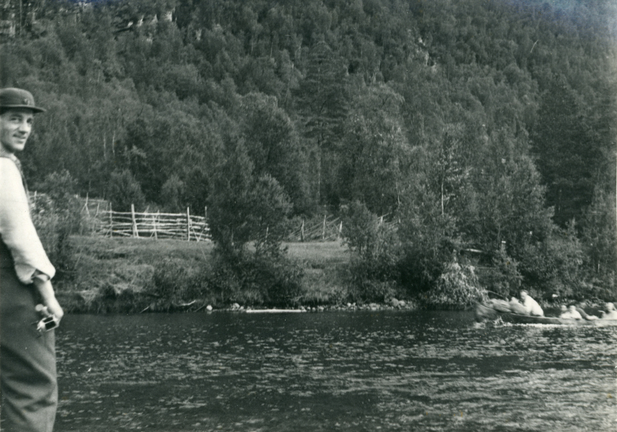 Mann med fiskestang ved Reisaelva. I bakgrunnen sees en elvebåt med følge på tur ned elva.