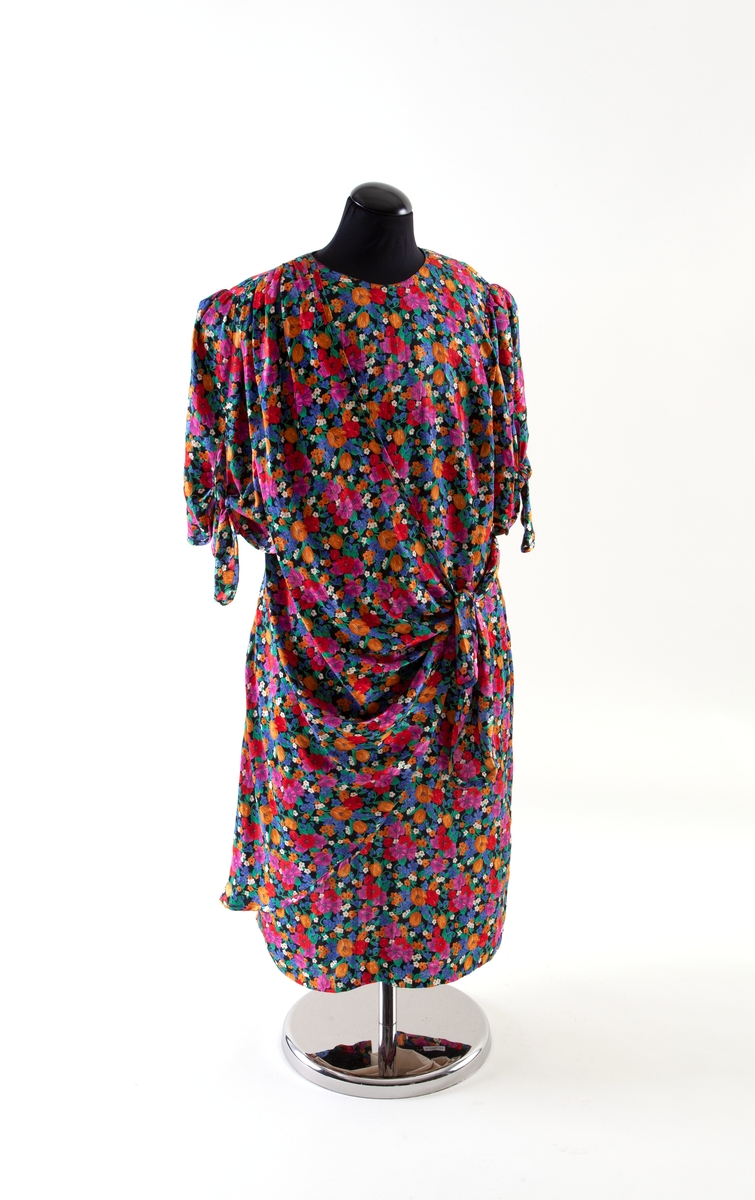 Blomstermønstret, flerfarget kjole med drapert stoff knyttet i et belte foran.