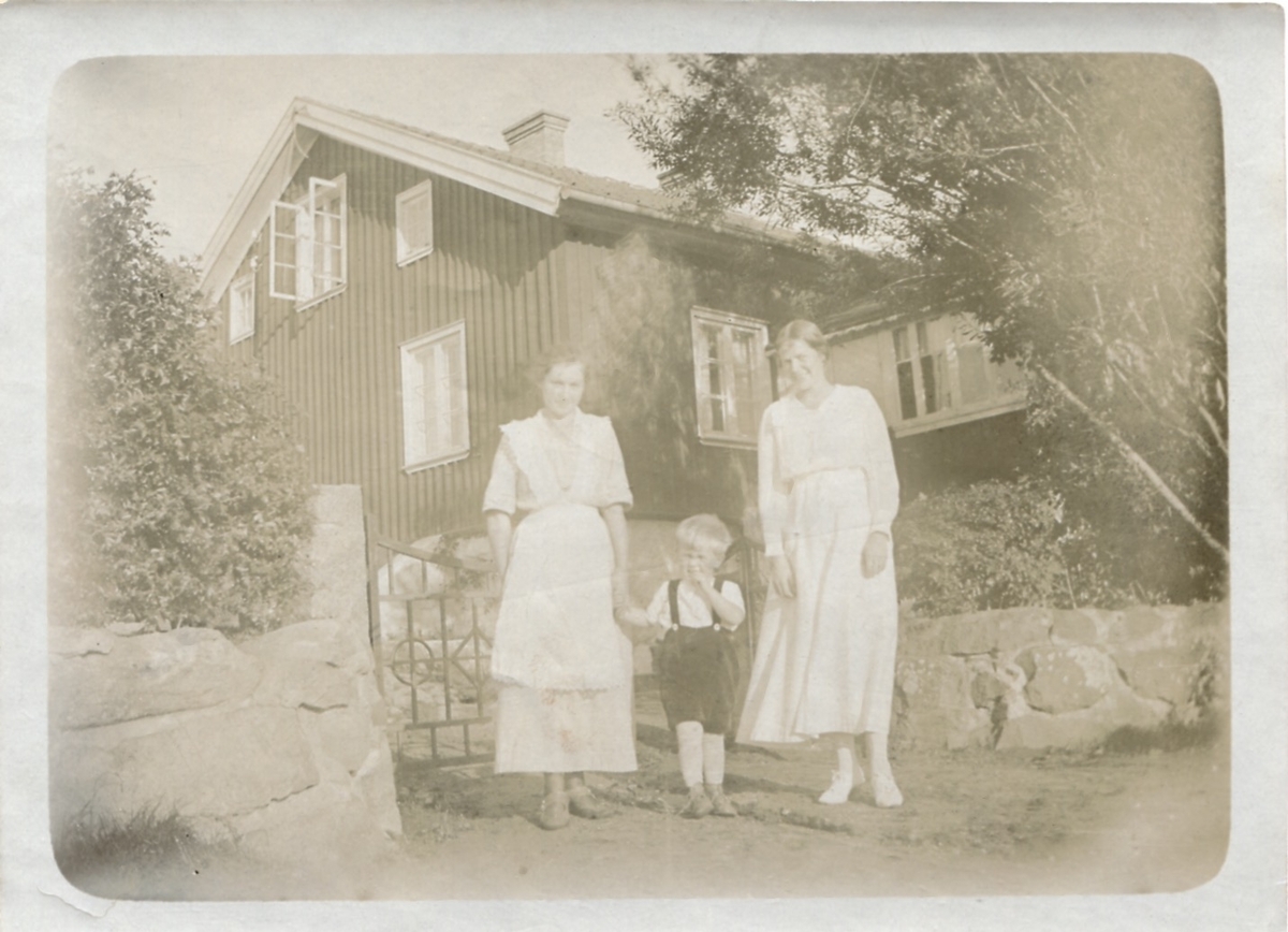 Två kvinnor och en liten gosse står utanför en stenmur. Ett hus ses i bakgrunden, okänd plats och årtal. Foto från Sagered-album.