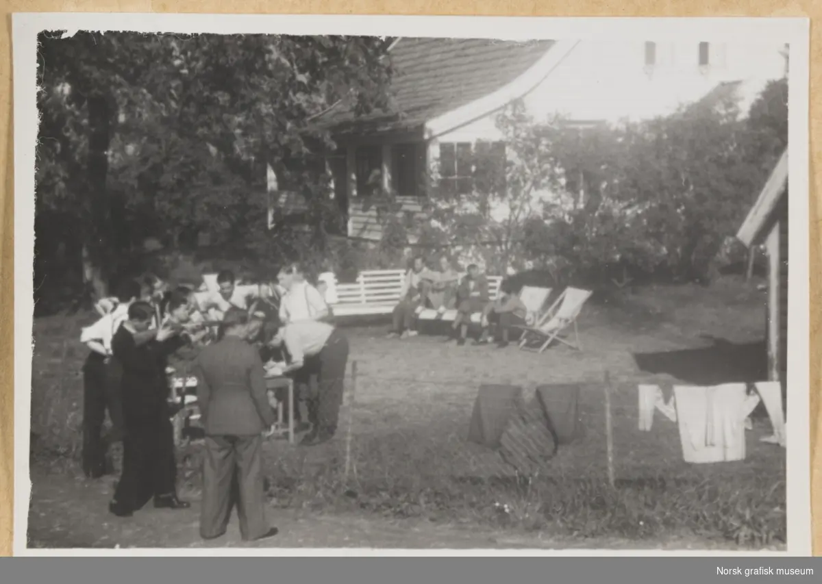 Utendørs, i en hage foran et hvitt hus. I forgrunnen står to menn foran et nettinggjerde som det henger noen klesplagg på (den ene sikter på noe utenfor bildet med et skytevåpen). I mellomgrunnen sitter mennesker på benker og stoler. Fotografert i forbindelse med Vestlandsk Trykkerstevne i Bergen, 1946.