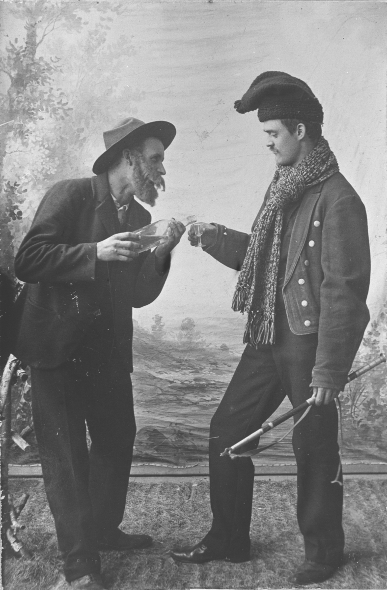 Atelierfotografi av to menn, utkledd i folkelige drakter. Angivelig scene fra et skuespill. Ukjent identitet og sted, men trolig rundt 1910.