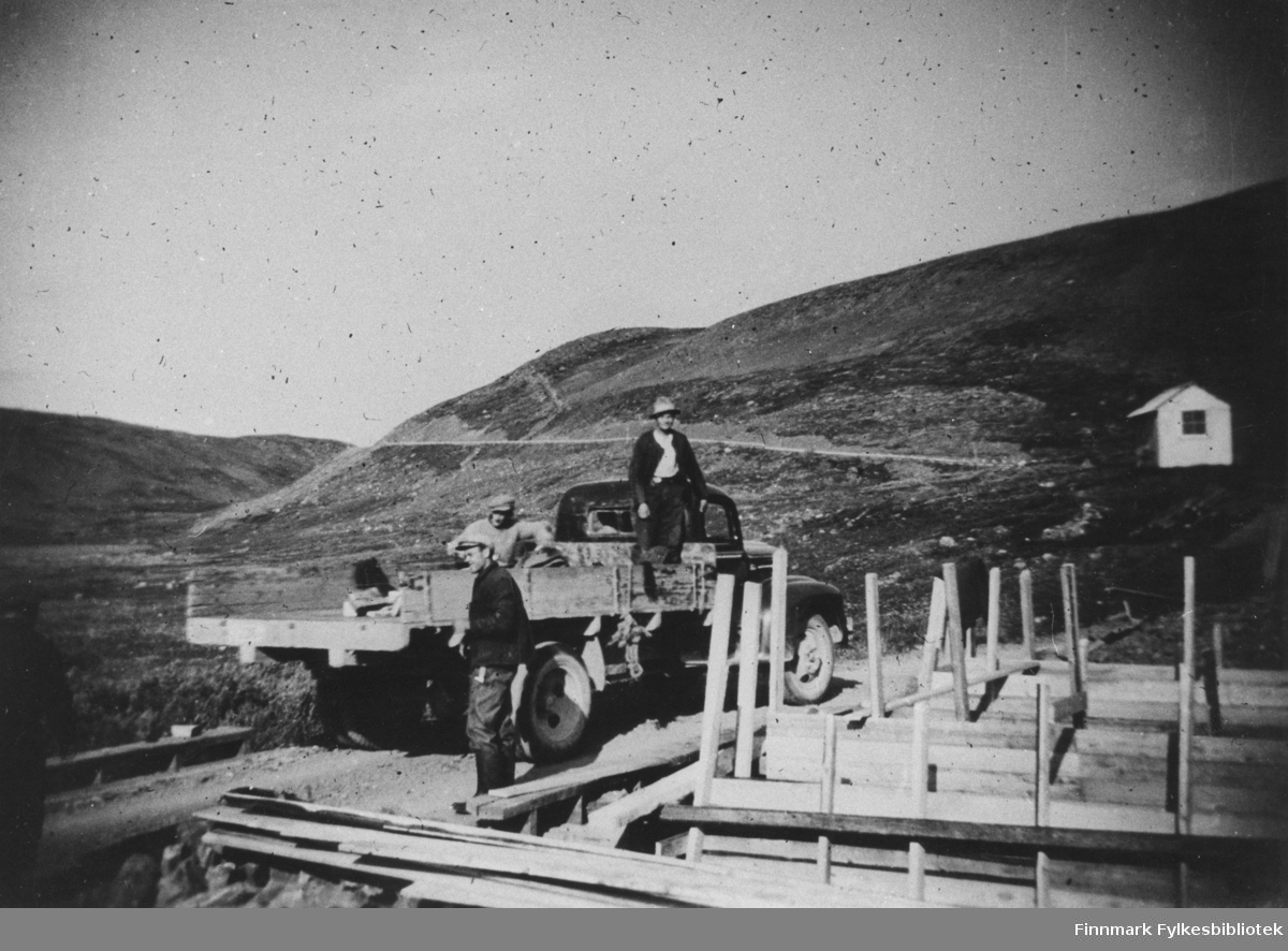 Krigskadeutbedringer i Finnmark 1946. Bruarbeide, provisorisk bru. På bildet ser man to menn som er på lasteplanet til en lastebil og en mann står ved siden av. Til høyre ser man den påbegynte bruen. Helt bakerst til høyre er et hus eller en brakke.