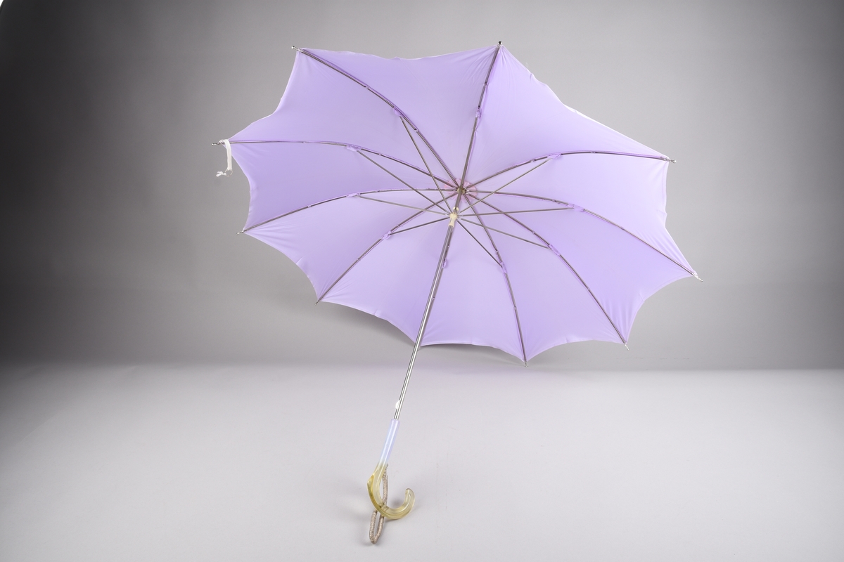 Paraply med 10 spiler og stang i metall. Handtak i plast, tofarga, lilla og gulkvit gjennomsiktig. Hempe i tekstil for å ha handa i. Lys lilla duk i kunstfiber.