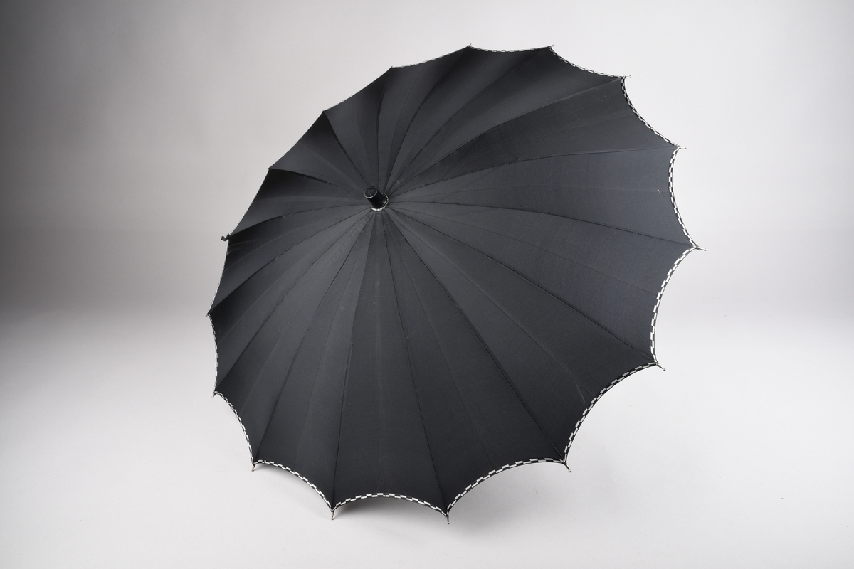 Paraply med 16 metallspilar. Stong i tre. Trekt med stoff i kunstfiber, ensfarga svart med dekor i sølv/svart rutemønster. Handtak i plast med hempe i tekstil til å ta inn på handa. Hempe for lukking av paraplyen.