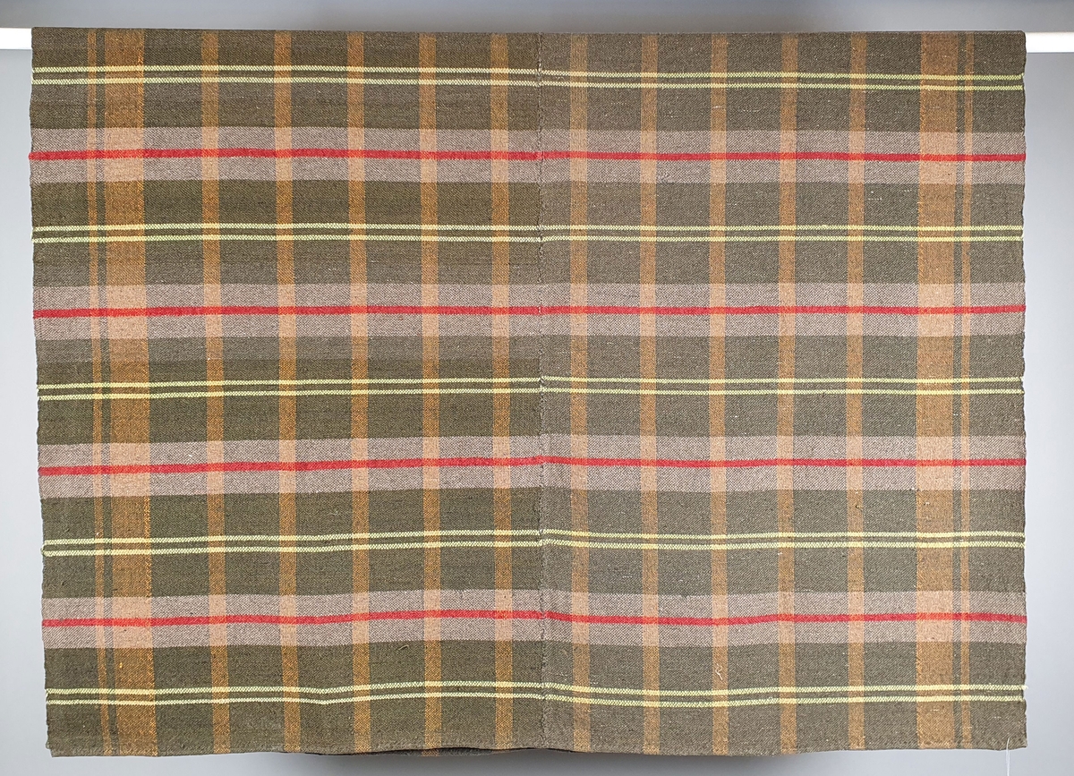 Grønt ullteppe med ruter og striper i rødt, gult og hvitt. Teppet er sydd sammen av to like deler.