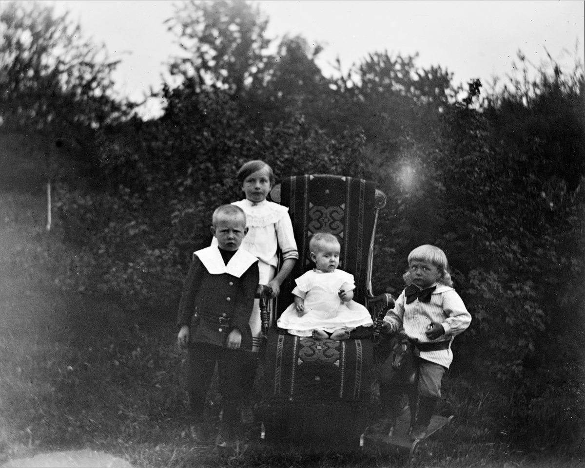 Fyra finklädda barn är samlade för fotografering utomhus. Den minsta sitter i en gungstol draperad med en vävd textil och gossen till höger sitter på en hjulförsedd trähäst.