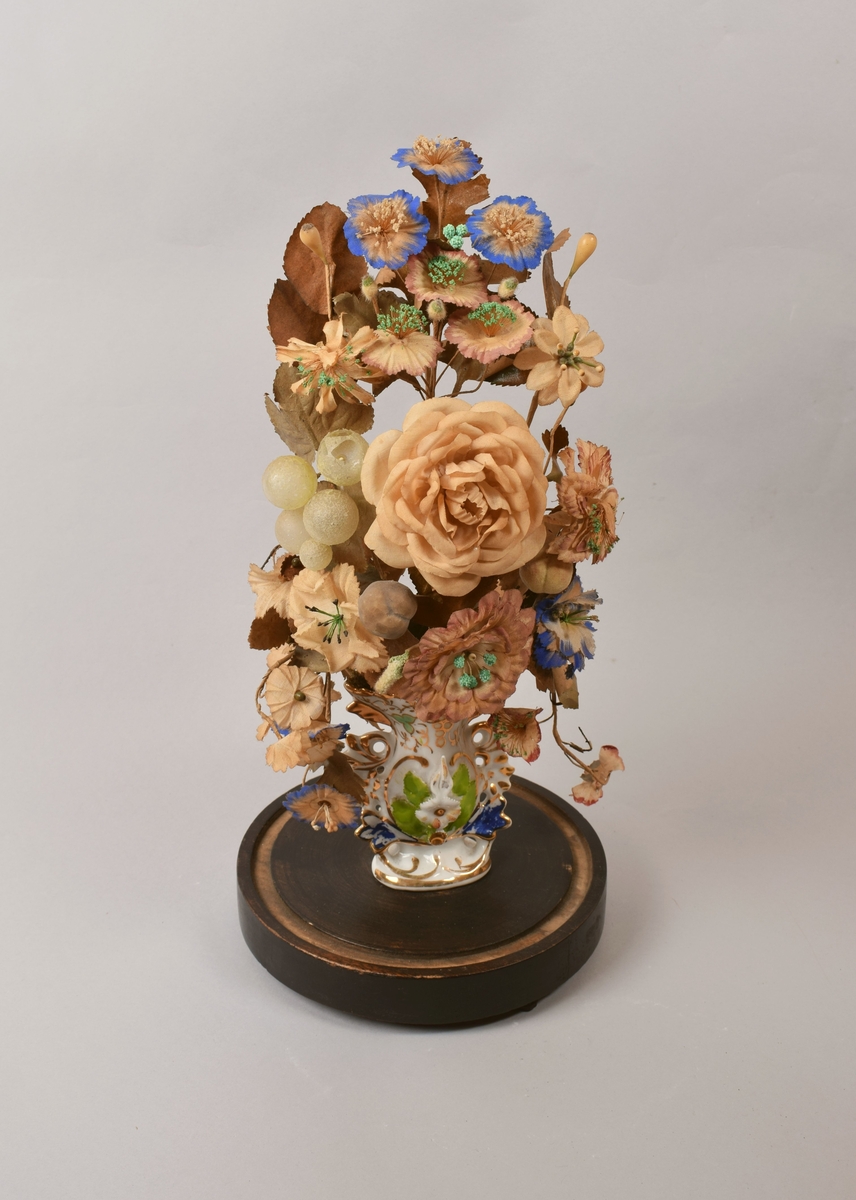Oppsats med kunstige blomster og frukt i liten porselensvase. Vasen med bladmotiv og forgylling på forsiden. Hele oppsatsen dekket med en glasskuppel.