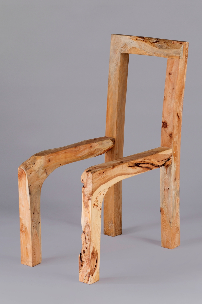Skulpturelt objekt i tre, utformet som en stol uten sete. Høyreist rygg som består av to stolper som glir over i bakbeina, og forbundet hverandre med et toppstykke. Sarg og forbein er laget av samme treemne.