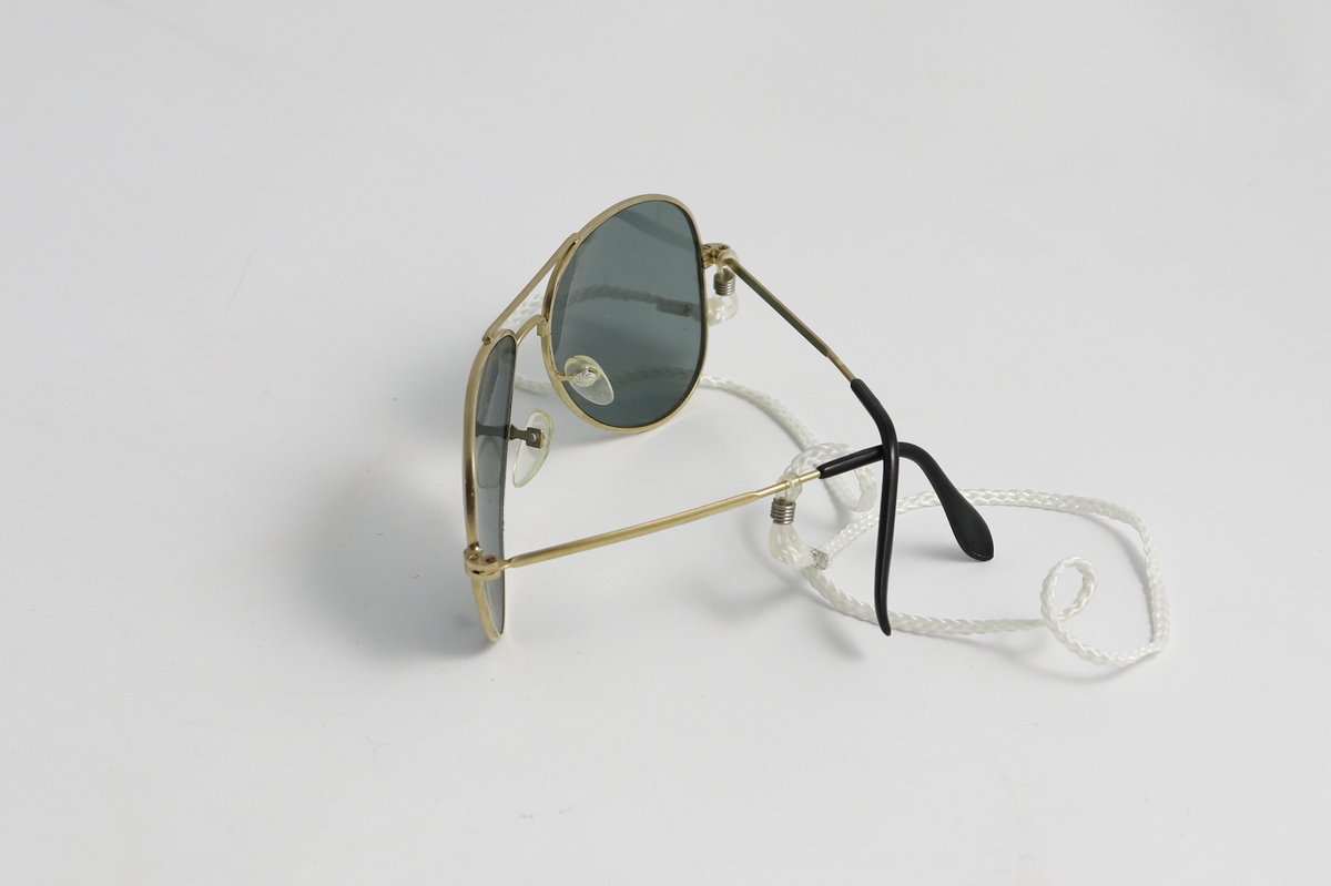 Pilotbriller med mørke glass og gullfarget metallinnfatning. Svarte plastikk endestykker på innfatning. Lite tau er festet på gjesntanden