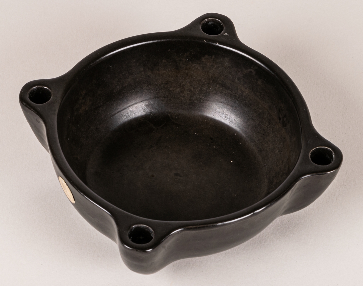 Ljushållare i form av en skål med plats för mindre ljus i varje hörn, lergods, svart glasyr. Eventuellt ursprungligen formgiven av Allan Ebeling 1924-1928, men pga etikettens utseende är den troligen tillverkad under 1950-60-talen.