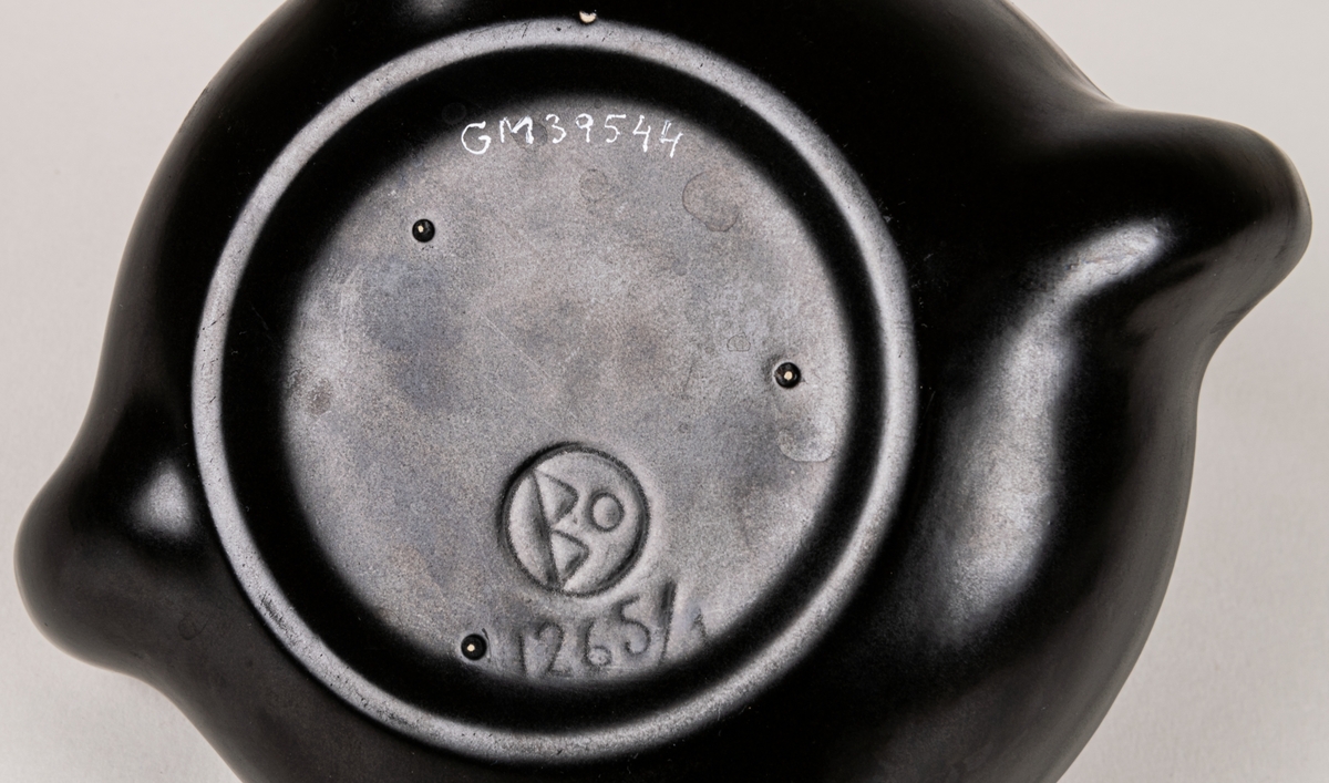 Ljushållare i form av en skål med plats för mindre ljus i varje hörn, lergods, svart glasyr. Eventuellt ursprungligen formgiven av Allan Ebeling 1924-1928, men pga etikettens utseende är den troligen tillverkad under 1950-60-talen.