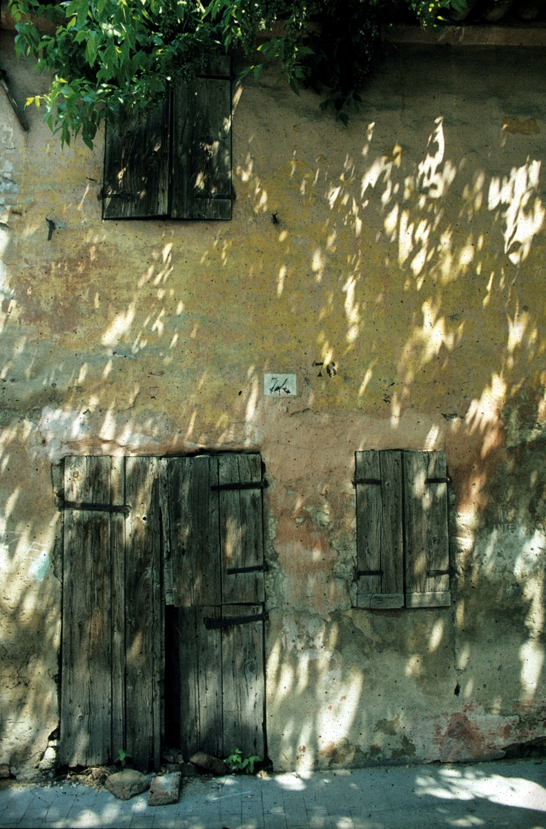 Foto av en vegg med dører og vinduer lukket med lemmer. Sola titter gjennom bladverket og kaster lysflekker på veggen.