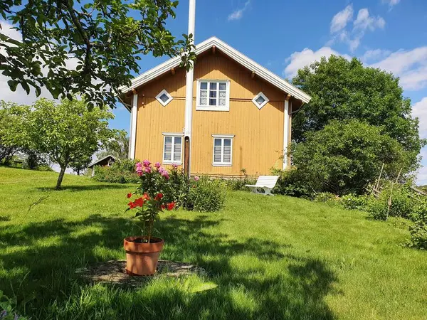 Hagen på Almenninga Eidskog bygdetun. Gult hus i bakgrunn. Flaggstang. Epletrær og andre busker. Hvit hagebenk.