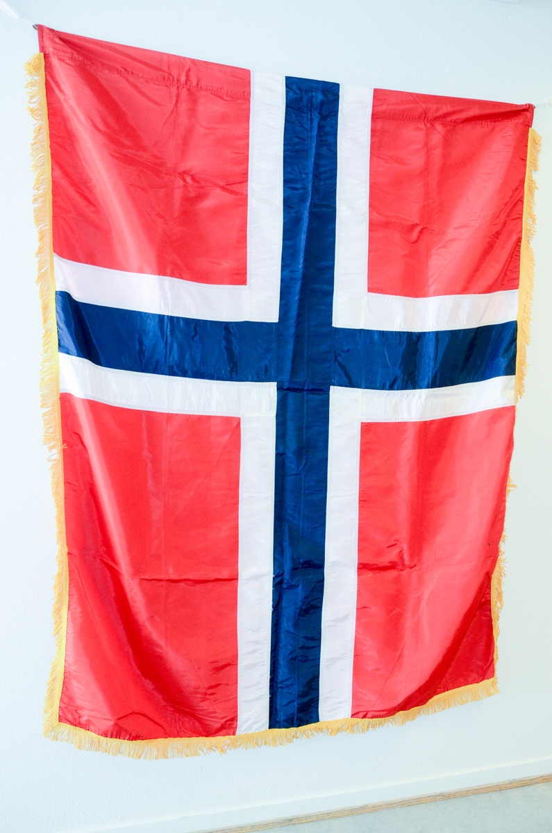 Det norske flagget med gule tvinnede frynser rundt kanten. Maskinsydd. Kunstsilke. Løpegang i den ene tverrenden, foret med linstoff.