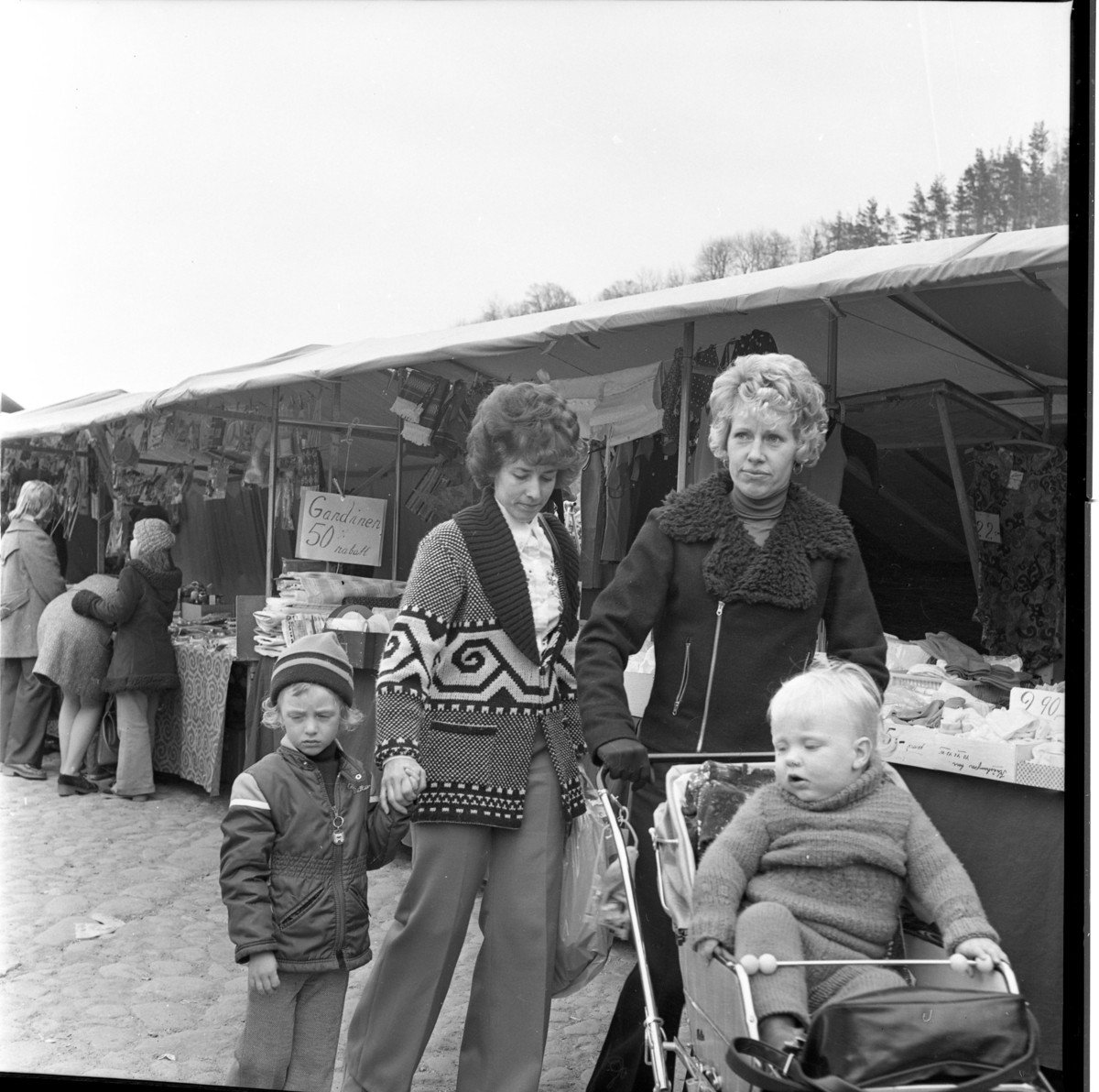 Två kvinnor går utmed marknadstält på torget i Gränna. Den ena kvinnan kör en sittvagn med ett barn och den andra kvinnan håller ett barn i handen.