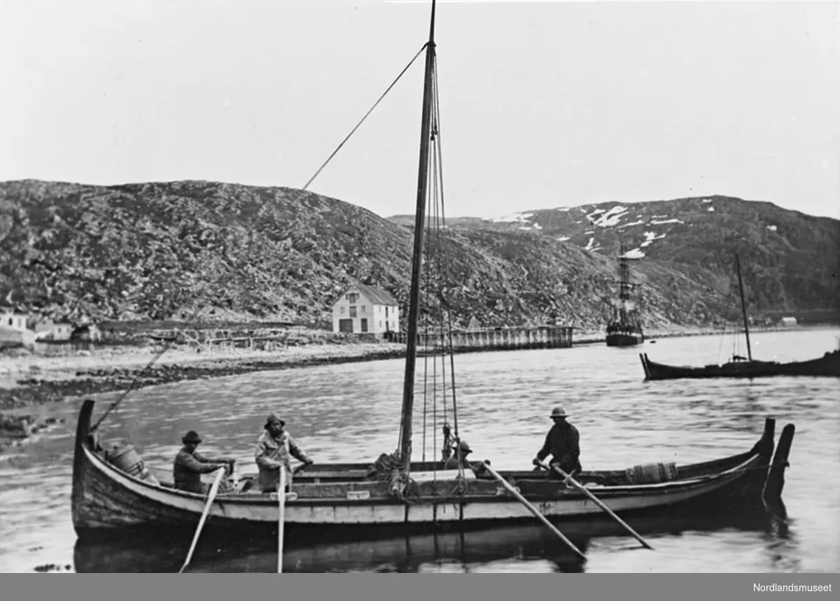 Nordlandsbåt, åttring med lauparrigg, 4 menn ror. I bakgrunnen en kai og en jekt til høyre.