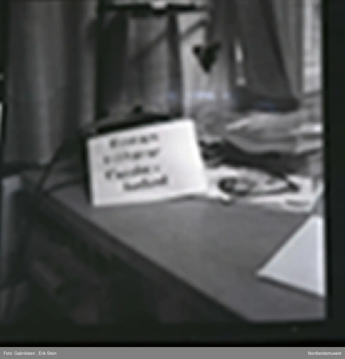 Fotografi av en lapp som står på en kontorpult til et kontor 
over en svart telefon som det går noen ledinger til 
og noen kasser med noen bøker og et avisutklipp og et kort med et klovneansikt 
og noen papirer og aviser som ligger på pulten
som man ser står vedsiden av to vinduer 
med gardiner og man ser en lampe med en lyspære
over pulten og man ser en åpen skuffe på pulten