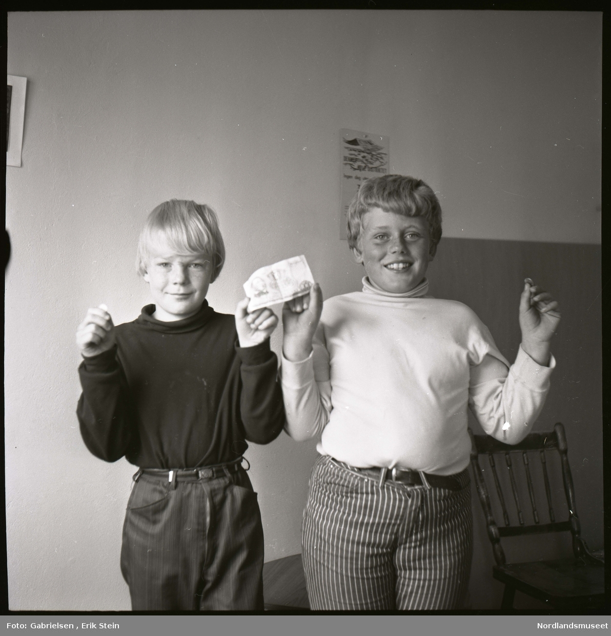 Fotografi av to gutter kledd i svarte og hvite skjorter som 
og stripete bukser som står i et rom foran en vegg som det henger en kalender og et bilde og en stol som står vedsiden av veggen og holder en pengeseddel