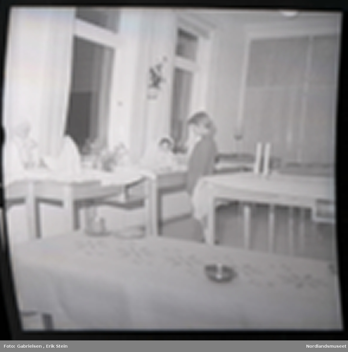 Fotografi av et barn som står inne i et rom og ser på en dukke
som ligger på en bord med en hvit duk 
som står mellom fire bord som står vedsiden av vinduer med gardiner som det ligger hvite duker på og en dukke som står lent på en vegg mellom to vinduer og man ser to andre bord
som står i rommet med duker på og to stearinlys som står
på bordet som står bak barnet i rommet og man ser
en potteplante som henger på veggen mellom to vinduer