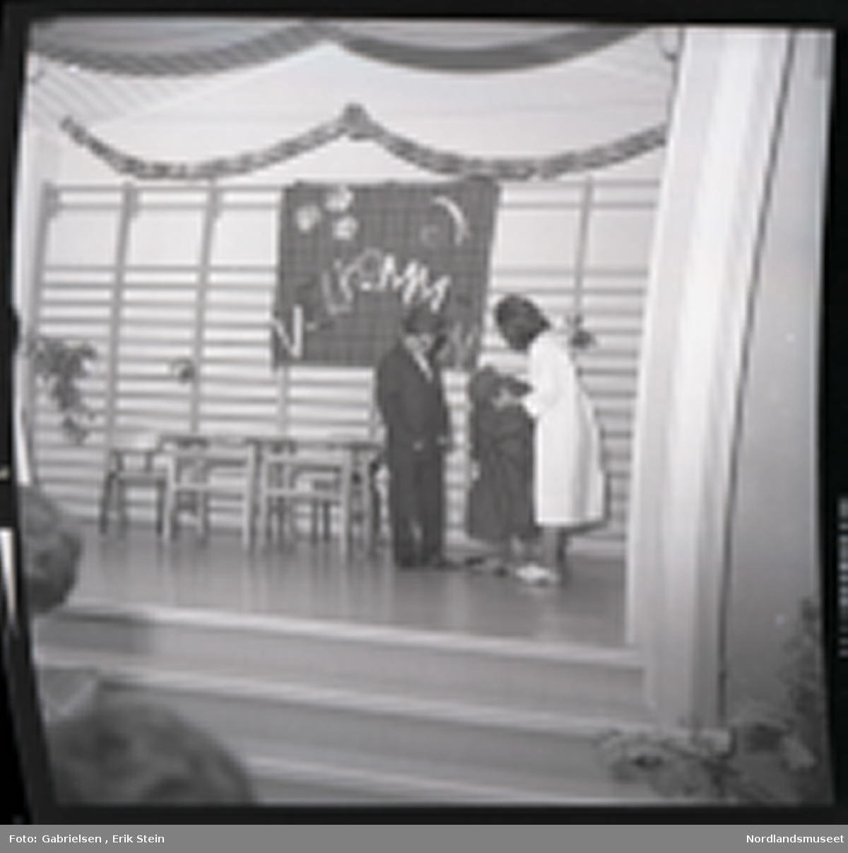Fotografi av en gutt og en jente kledd i en dress
og en hvit skjorte som står på et scene med klatrevegger
og to trappetrinn som går opp til scenen og to stoler 
og et bord som står på scenen og noe pynt 
som henger over klatreveggen og en duk som henger
på klatre veggen som står velkommen på 
man ser at jenten holder et svart klede mens gutten 
står og ser på mens publikum sitter på stoler i salen
og ser på forestillingen