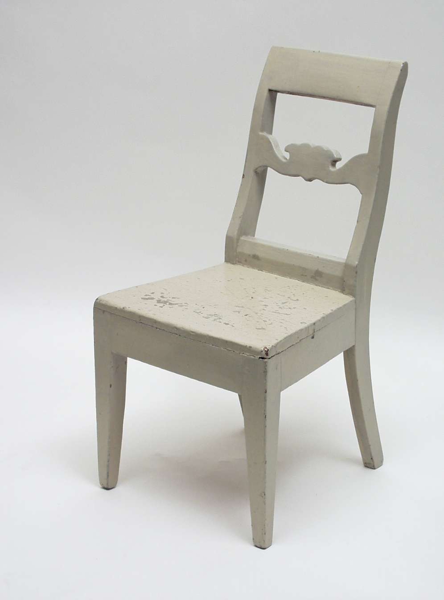 Liten gråmalt stol i gran med utskåret ryggstykke. Stolen har glatt skulderstykke og firkantede ben - forben rette, svungne bak. Setet smalner bakover.