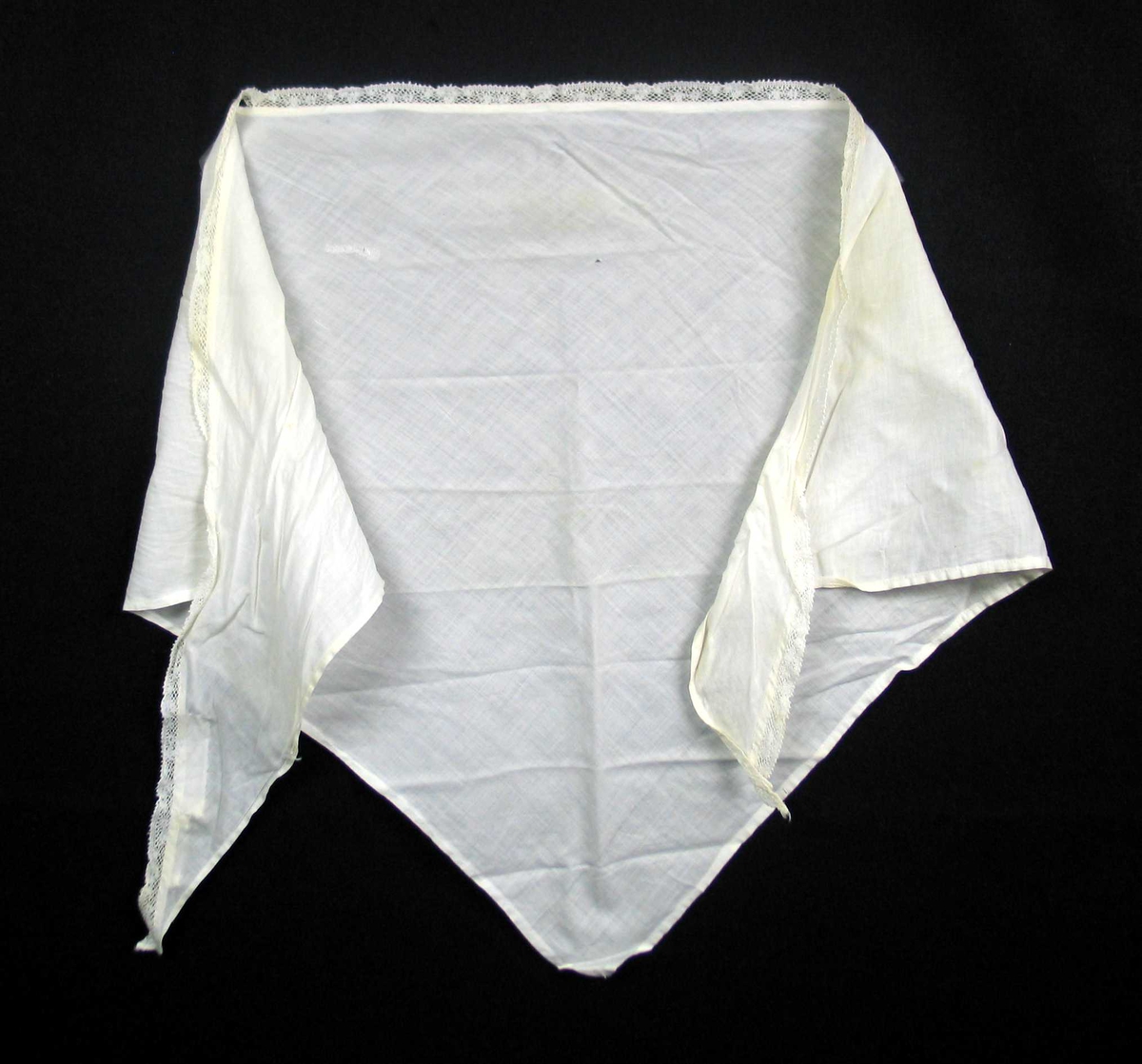 Hvitt trekantet tørkle i to-skaft vevd bomull med påsydd kniplingsblonde. Blonden er antatt maskinvevd. Tørkledet er maskinfaldet på tre sider