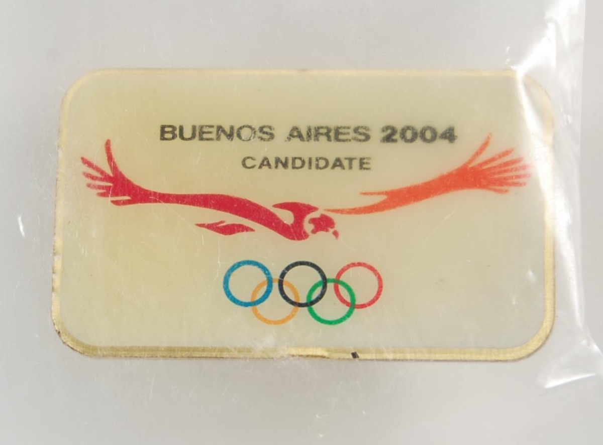 Hvit pins med logo for kandidatbyen, Buenos Aires, i forbindelse med søkerprosessen til de olympiske leker i 2004. Logoen består av OL-ringene i farger og en rød og oransje fugl, som antakeligvis er en condor.