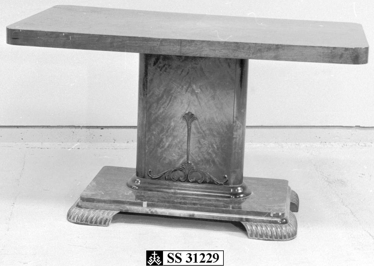 Et bord i furu som er lakkert i brunfarge.
Det har firkantet midtsøyle med utskjæring i form av blomst. Midtsøylen står på en plate med fire ben.

