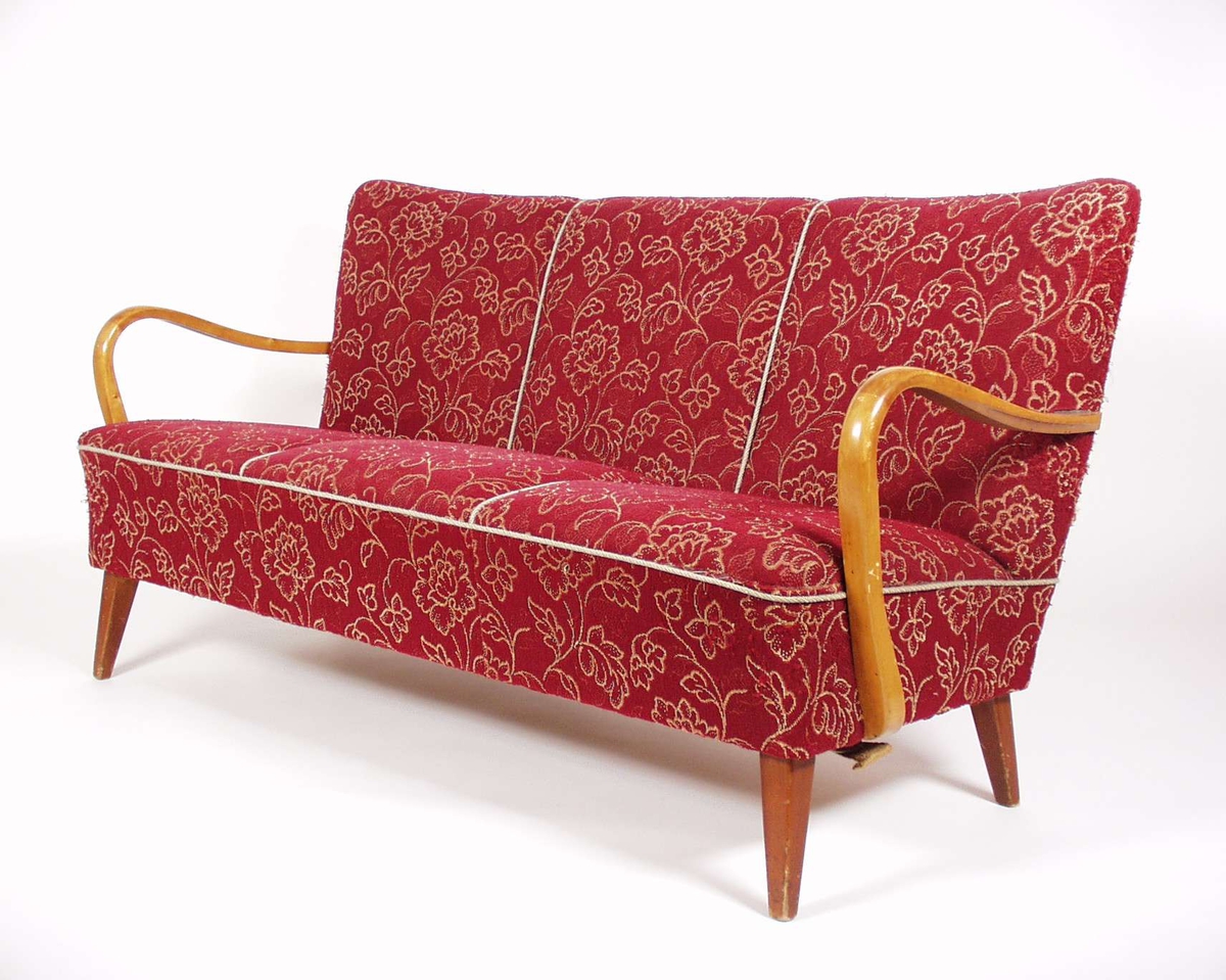 Sofaen er stoppet med rødt blomstermønstret møbelstoff, armlenene og beina er i lakkert bjørk.