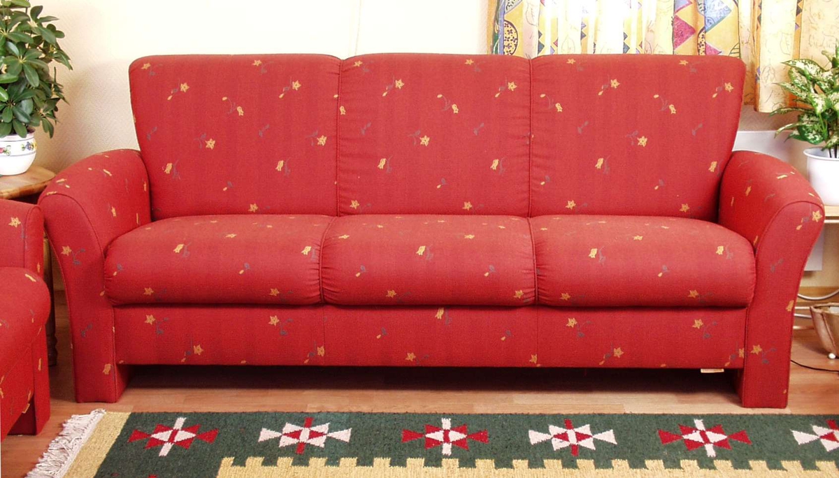 Sofa heltrukket i rødt stoff med gule blomster og grønne blad. Armlenene er buede og ryggen skråner lett utover. Sete og rygg har stikninger som deler sofaen i tre.


