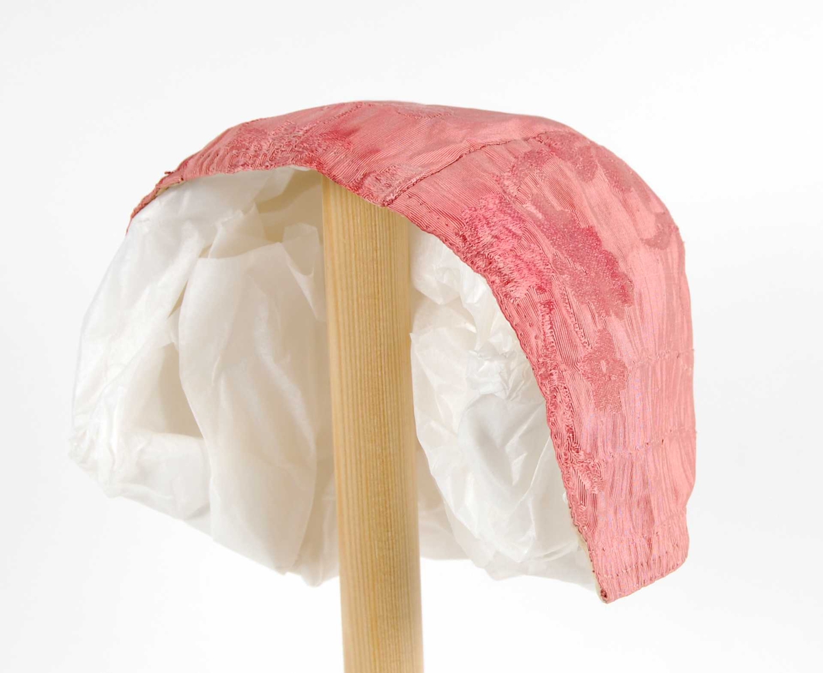 Rosa mønstervevet silkelue med fôr av bomull. Et stykke fra panne til nakke, et stykke som er avrundet mot midtstykket på hver side.