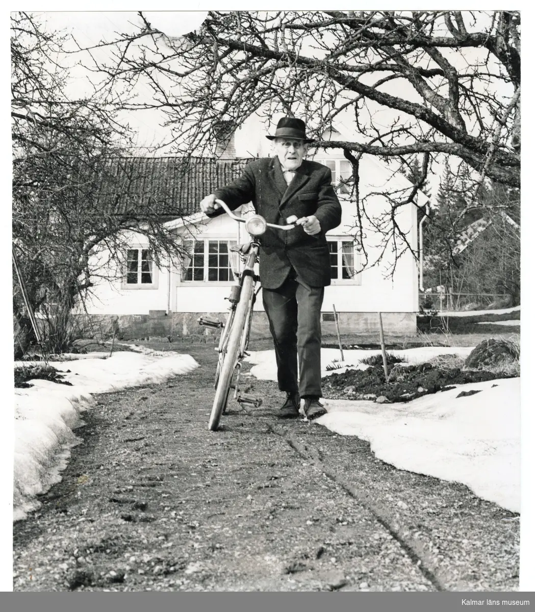 Gunnar Edvin Franzén gående med sin cykel framför huset på gården Skogstorp, Tomtabyholm omkring 1970. Påskrift baksida: Gunnar Franzén omkr. 1970.