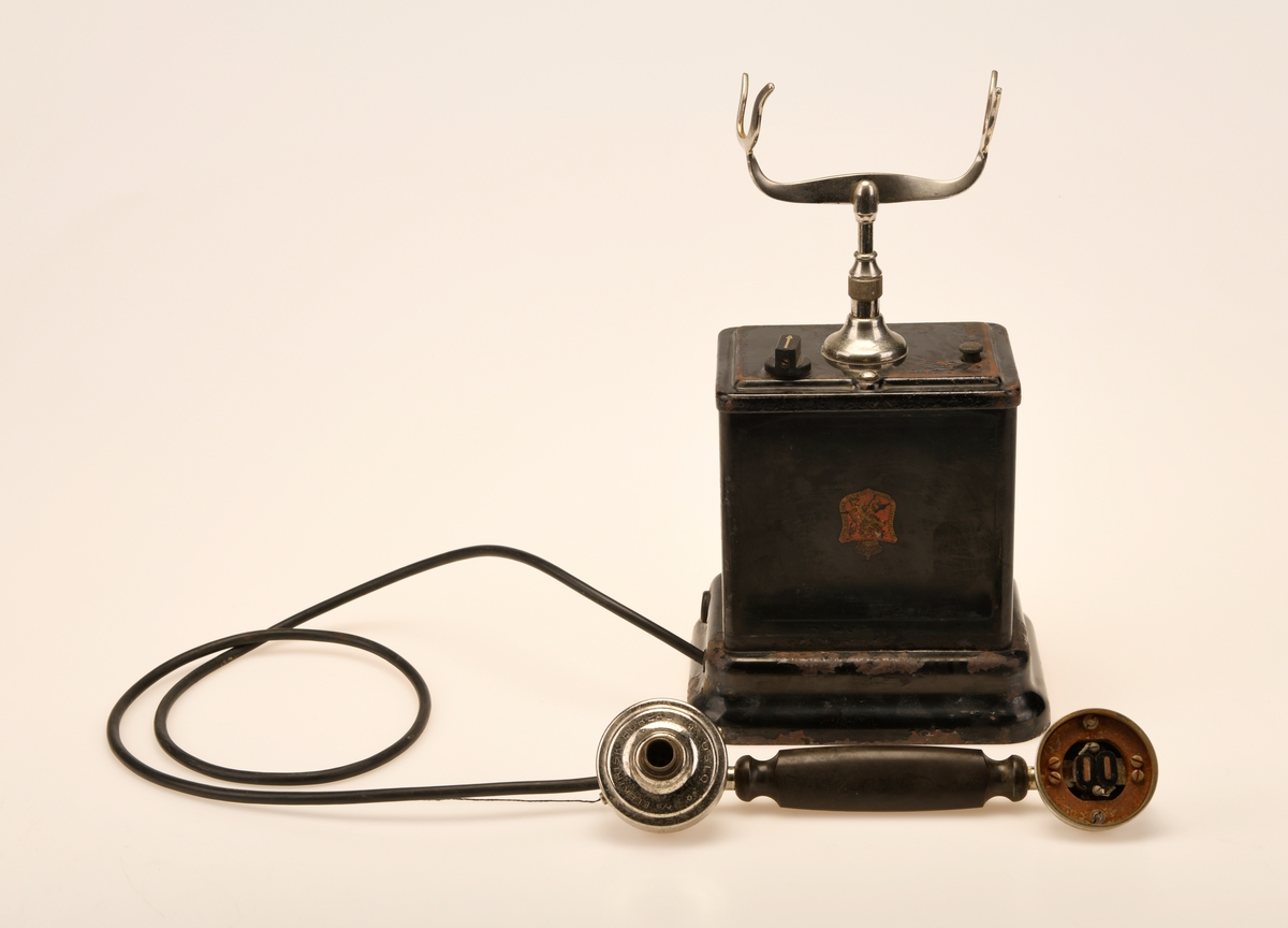 En svart bordtelefon av Elektrisk Bureau sin 1922 modell. "Den norske løve" pryder forsiden og baksiden av aparatkassen, disse er slitt og i tillegg oppned. Forøvrig har telefonen en y-gaffel. Ledningen som går fra aparatkassen til telefonrøret er av rund plast, noe som tyder på at den har blitt byttet ut på et tidspunkt. Sveiven mangler, deler av telefonrøret mangler, og ledningen med støpsel er fjernet.