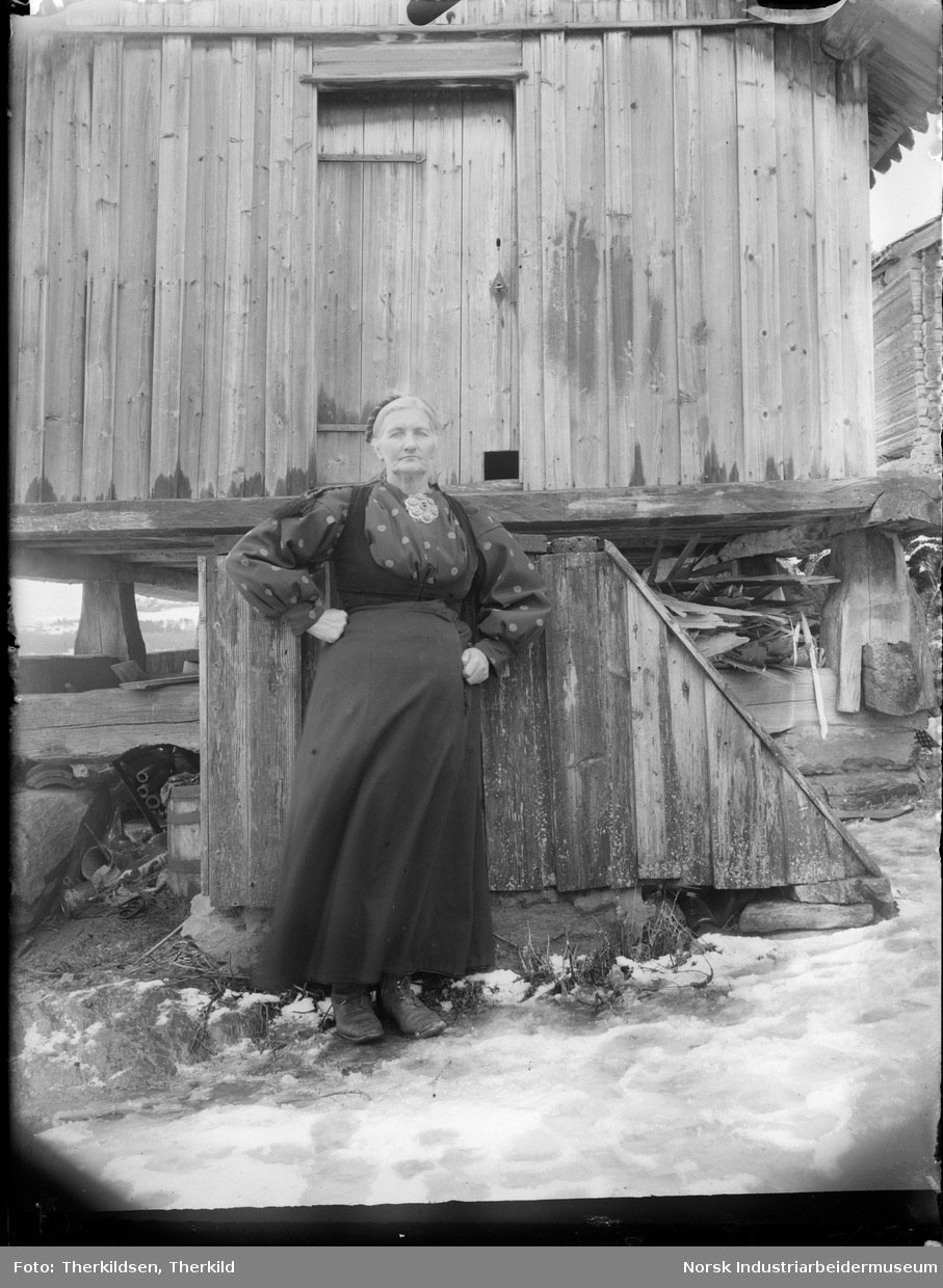 Kvinne i stakk og liv med sølje i halsen stående foran bur på den gamle husmannsplassen Merde under Nordre Tinnes.