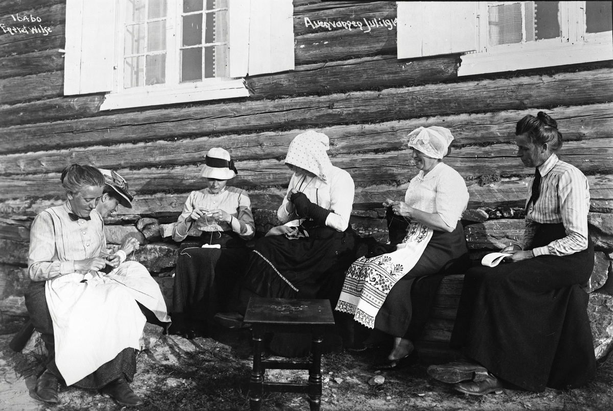 Kvinner sitter i solveggen på ei tømmerhytte og strikker ved Auenvangen, Gol, 1914.