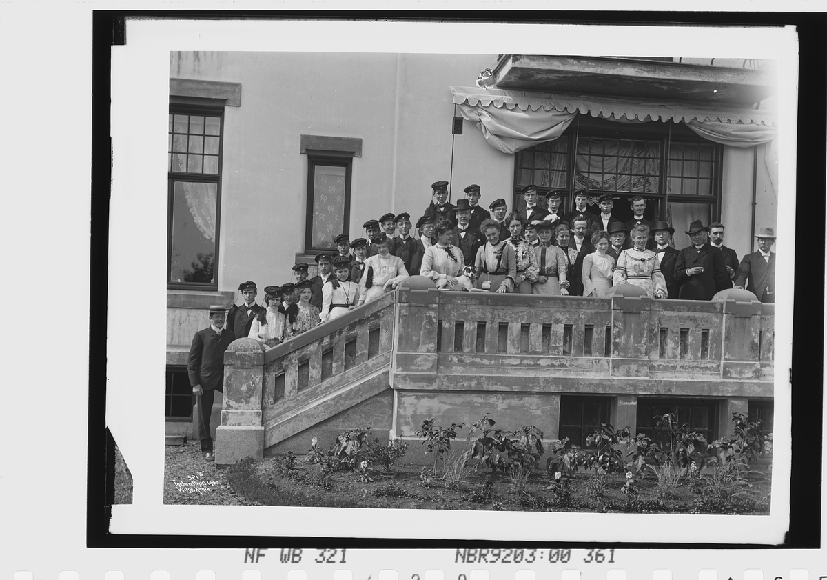 Skoleavslutning, avgangsklasse studenter med studentluer samlet på verandaen, skolebestyrer Andersen nederst i trappen.