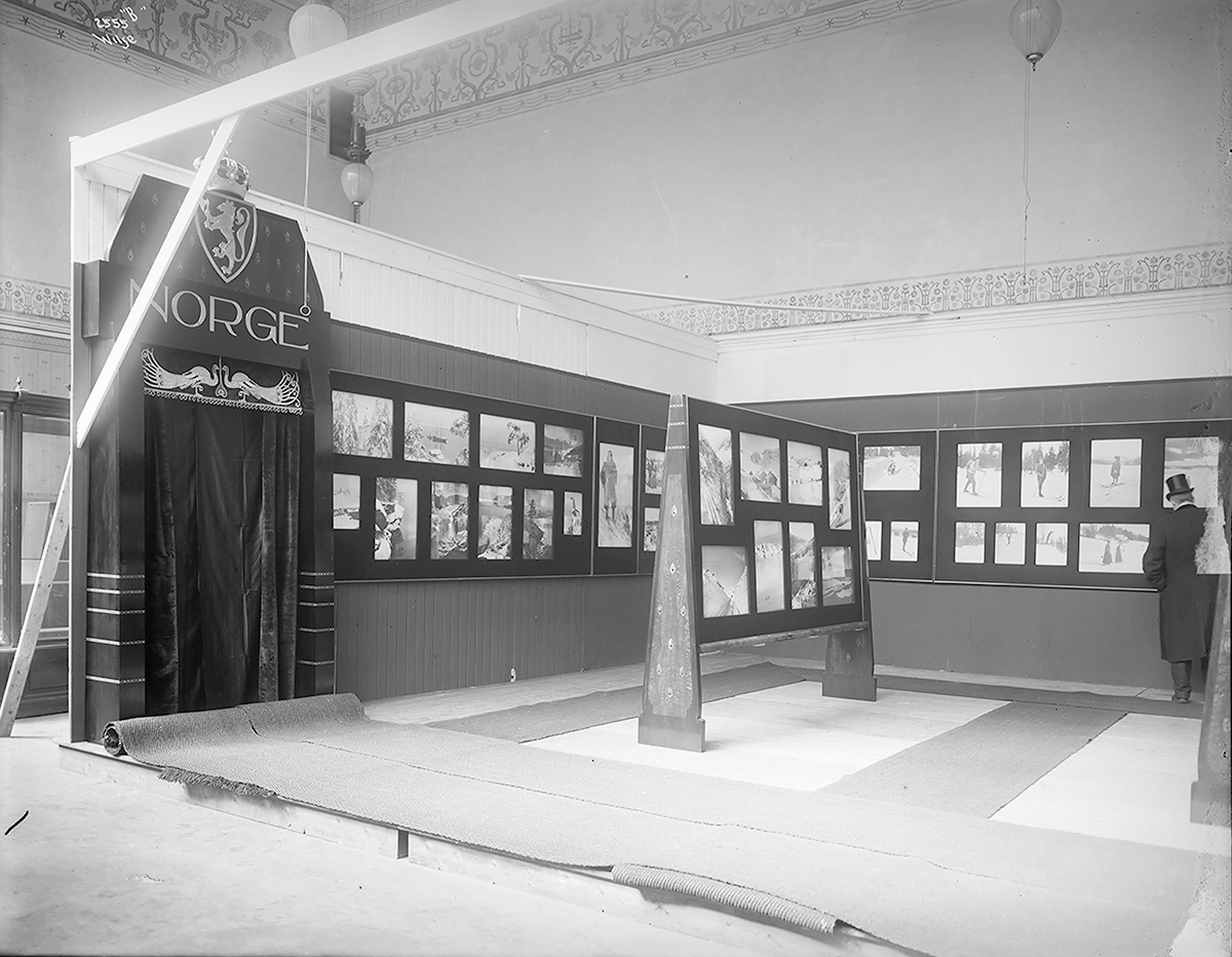 Før åpning av utstilling med Wilse-fotografier i Dresden. Til venstre døråpning med portierer under skilt med riksløven, teksten "Norge" og dekor med påfugler. Fotografert 1909.