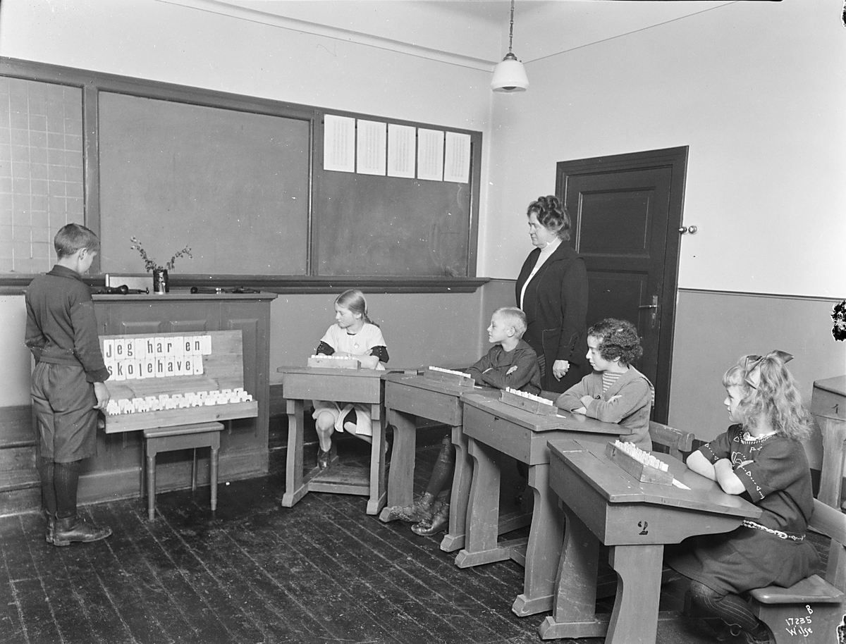 Elver sitter ved pultene sine og ser på en gutt setter opp setning på en tavle. Lærerinne Jacobine Ulrica Rye står i bakgrunn, Granhaug offentlig skole. Fotografert 1923.