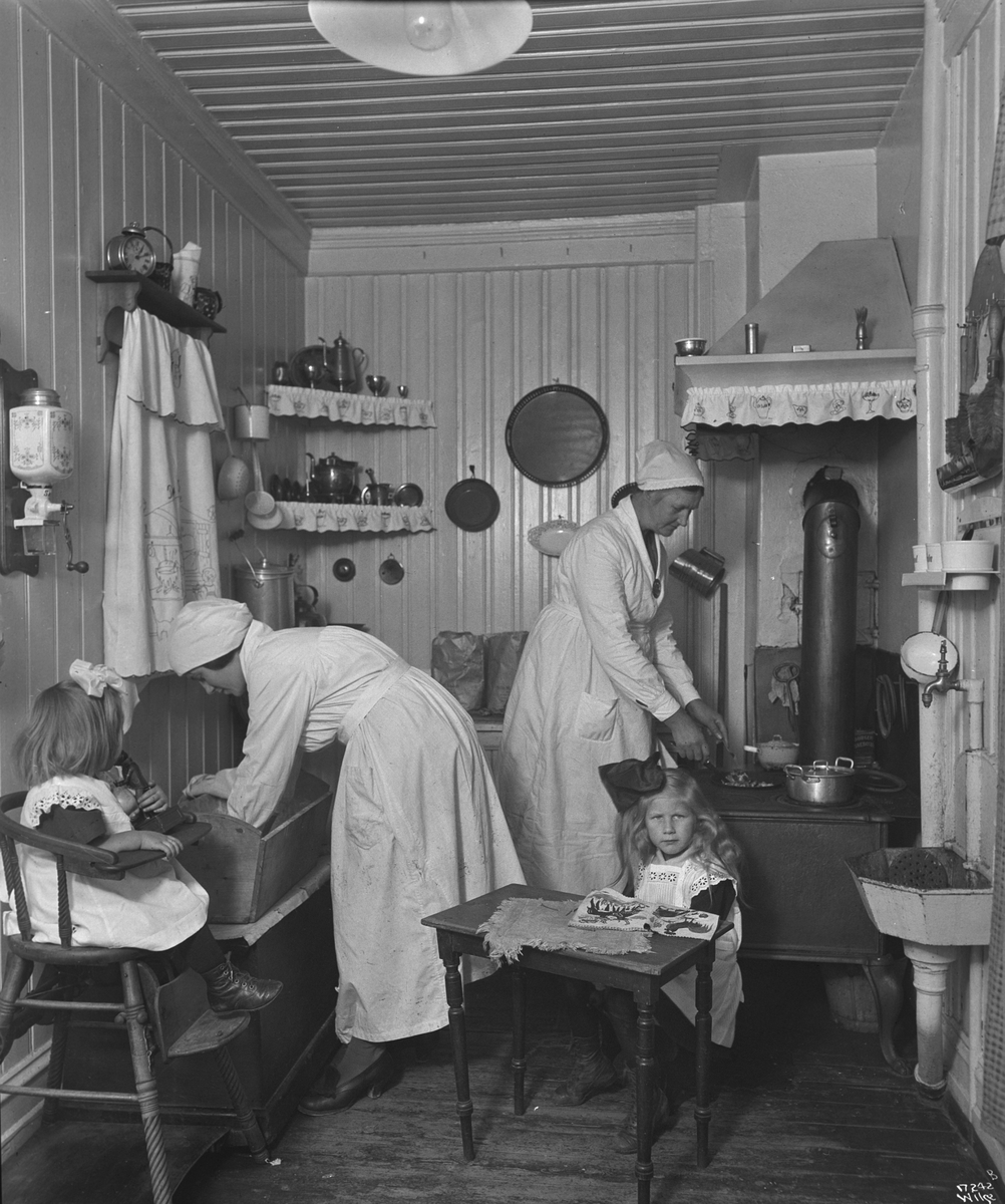 Hjemmekonsulenter steller på et kjøkken, to små piker sitter på stoler. Fotografert 1923.