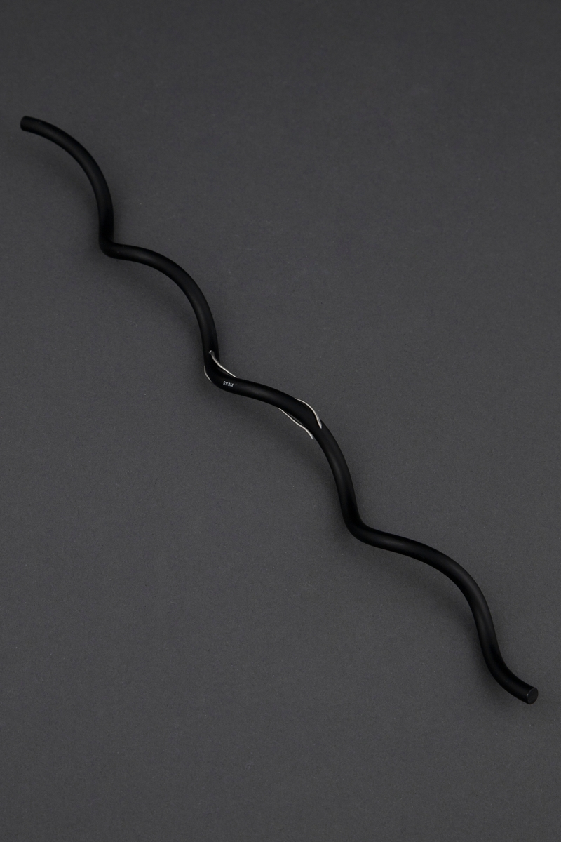 Brosje i form av en bølget, svart aluminiumsstav. Gjennom denne er det stukket en dobbel, tynn ståltråd som nål for å feste brosjen.