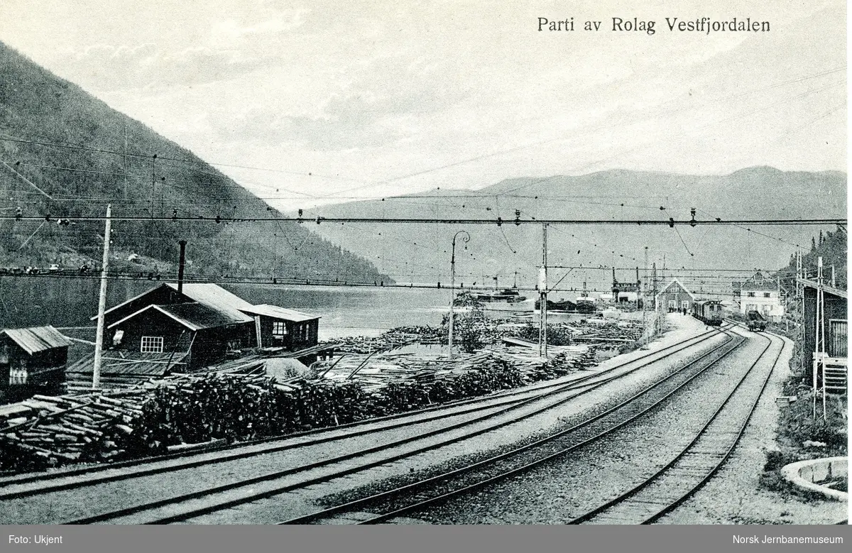 Mæl stasjon med store mengder tømmer, klart for fløting over Tinnsjø. I bakgrunnen elektrisk lokomotiv med tog til Rjukan. Ved fergeleier MB JARL, DS SKARSFOS og DF RJUKANFOS eller DF HYDRO