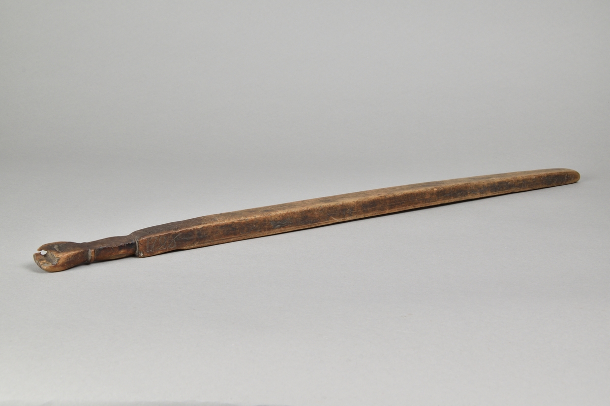 Träaln tillverkad i björk. Fyrsidig med rundat, nött avslut. Kort handtag, avslutat med snidad hand med hål för upphängning. Nedanför handtaget inskuret årtal, "1726".