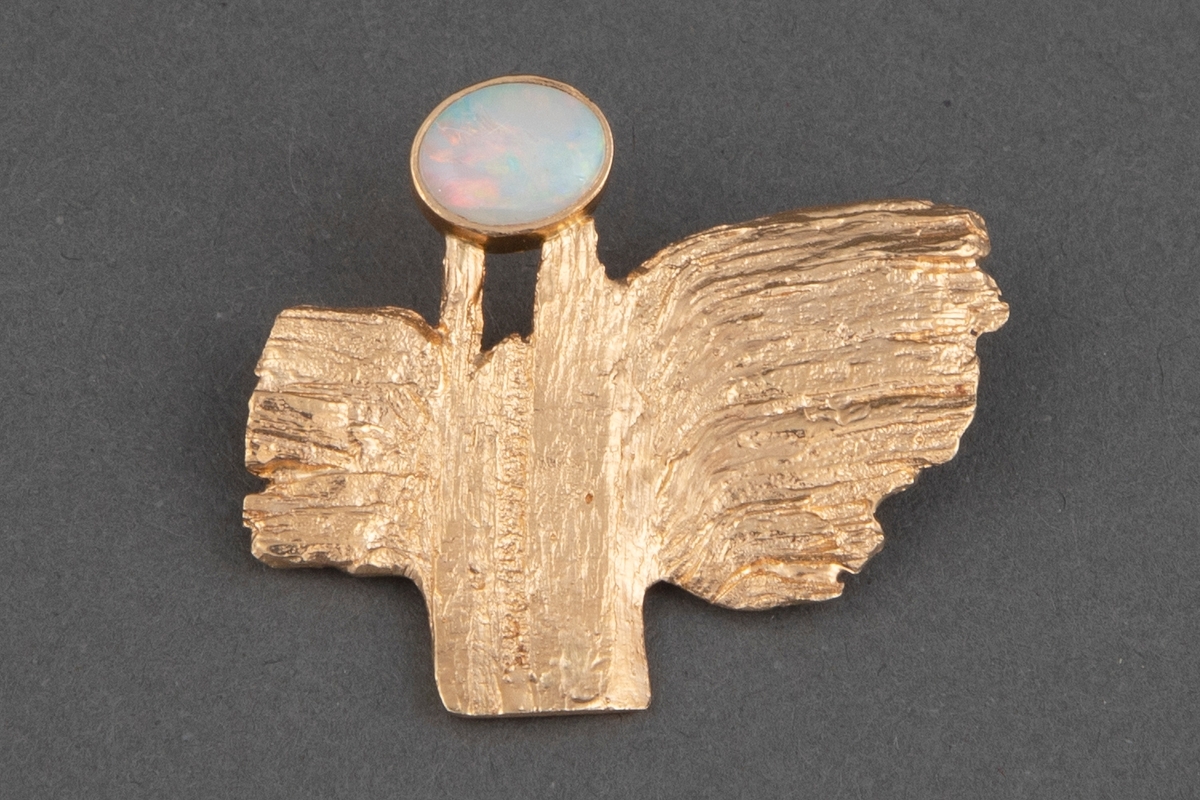 Brosje av gull i slyngestøpsteknikk med hvit opal. Brosjen har en uregelmessig form som minner om en fugl med vinger, stjert og hode. Overflaten er ujevn. Opalen er satt inn i en blank innfatning. Baksiden er glatt og har nål.