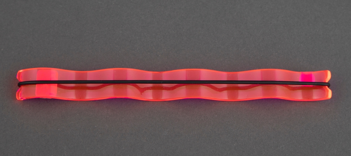 Brosje bestående av et avlangt, rektangulært stykke fosforiserende rød plast som er bøyd i bølgeform med fire "bølger". I hver kortside av plaststykket er det et lite innsnitt. En flat, svart gummistrikk er tredd rundt smykket og festet i innsnittene.