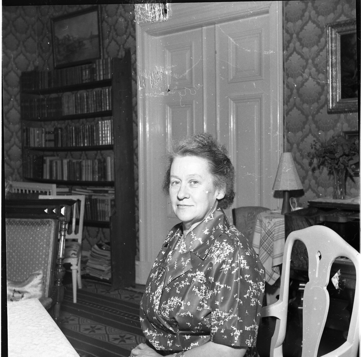 En kvinna sitter vid en matsalmöbel i en salong. I bakgrunden synsdubbeldörrar med speglar och bredvid dem står en bokhylla. Kvinnan har en kort frisyr och en mönstrad klänning