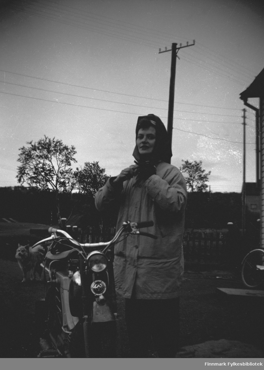 Kajsa Persen på moped utenfor bakeriet i Varangerbotn. Bak hunden Moen families hund Roy. Vegkrysset sees i bakgrunnen. Bildet tatt i 1957.