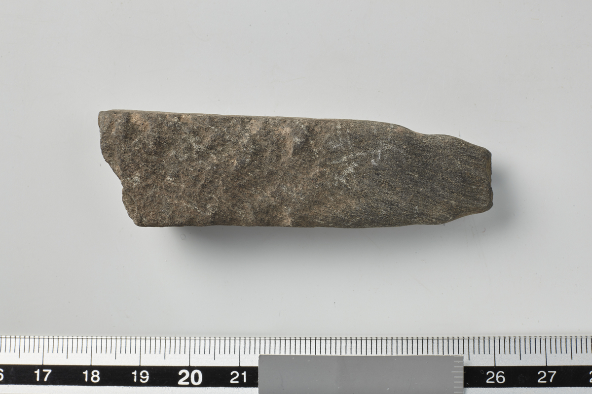 Avlangt, firkantet, flatt stenstykke tilspidset i den ene ende, hvor odden er avbrutt 7,7 cm l.